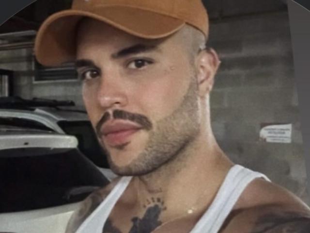 Photograph of MedellynDark with a light brown cap, white tank top, and tattoos on his neck and chest. He has a trimmed mustache, tattoos, and a cocky expression. Background includes a car.