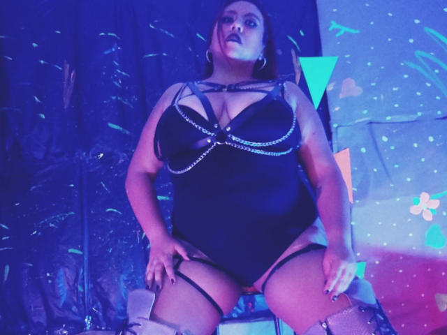 LexyDom stands in a low angle shot, wearing a black, strappy lingerie bodysuit with chain details, and thigh straps. She's illuminated by blue and pink lights, looking directly at the camera.