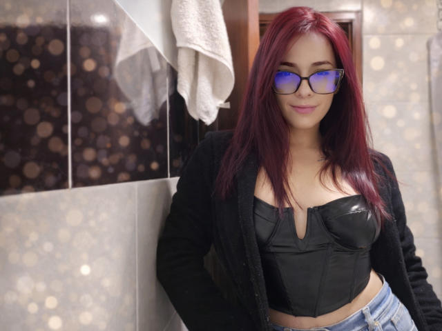 Photographie d'Ashley Platta avec des cheveux roux, des lunettes à monture bleue, un crop top en cuir noir, une veste noire et un jean bleu. Une serviette blanche est suspendue en arrière-plan. Pose sexy et confiante. Effet bokeh..