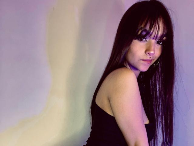 Photograph of MarietteJones, a sexy woman with long black hair and bangs, wearing a black strapless top, looking over her shoulder with a sultry expression, soft light, subtle shadows.