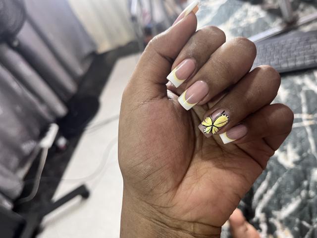 Close-up of a dark-skinned hand with long, pink and white French-tipped nails, featuring a small yellow flower design on the ring finger. Sunlight highlights the glossy nails, creating shadows.