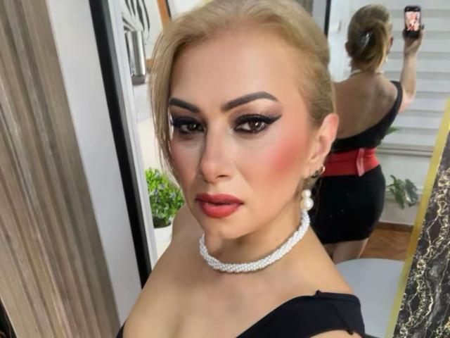 Photograph of MisseScarleth with blonde hair, heavy makeup, red lipstick, black dress, white choker, reflecting in mirror, showing back with red belt, sexual tension, seductive pose, confident expression.