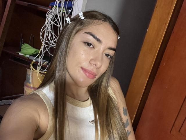 SharonKlart has long brown hair, wearing a white sleeveless top, with a shiny, slightly oily face. Her arm shows small tattoos. She's looking at the camera with a slight smile. Background includes wooden door and cluttered shelf.
