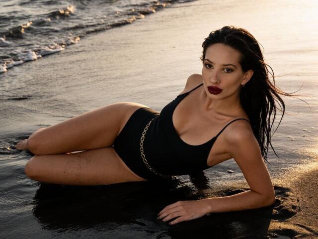 ElzaKomx giace su una spiaggia bagnata, indossando un costume intero nero con una cintura argentata, i capelli bagnati le aderiscono al viso. la luce del sole che le brillava sulla pelle, con le onde sullo sfondo.