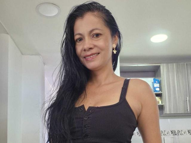 Photograph of MelissaBaud with long, wet black hair, smiling, wearing a black tank top with buttons, gold earrings, in a modern kitchen. Bright lights, white cabinets, gray wood. Fucking hot.