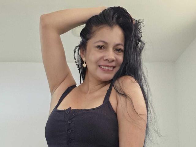 MelissaBaud, donna asiatica, capelli lunghi e neri, sorridente, indossa una canotta nera con bottoni, braccio alzato, mano tra i capelli, carnagione chiara. sfondo, leggera ombra a destra, casual, sicuro di sé, sexy, attraente.