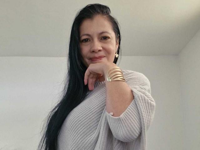 MelissaBaud, Asian woman with long black hair, white knit sweater, gold bracelets, leaning forward with hand under chin, smiling slightly, light skin, white background, subtle sexual tension, casual pose.