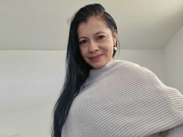 MelissaBaud, an Asian woman with long black hair, wearing a white knitted sweater, smiling slightly. Her hair partially covers her right breast, creating a subtle, sexy tension. Bright, minimalist background.