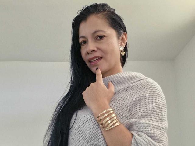 MelissaBaud, incarnato medio, capelli lunghi neri, maglione bianco in maglia, braccialetto dorato, orecchini dorati, dito sulle labbra, sorridente, leggermente inclinata la testa, luce naturale, posa informale, sottile tensione sessuale, espressione sicura..