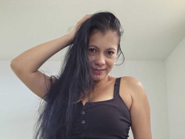 MelissaBaud with long black hair, smiling, hand in hair, wearing a black tank top with buttons, light skin, casual pose, slight sexual tension, indoor setting, soft natural light.