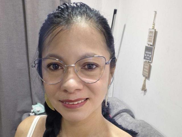 Primo piano di MelissaBaud, una donna asiatica con capelli scuri raccolti in una coda di cavallo, che indossa occhiali con montatura trasparente e rossetto rosso. Sta sorridendo, mostrando un po' di scollatura. Sfondo luminoso con cartelli..