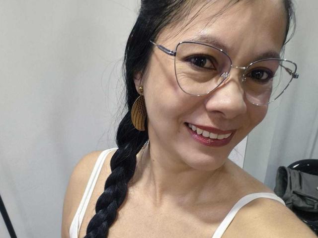 Gros plan de MelissaBaud, avec des cheveux noirs tressés, portant des lunettes transparentes et un débardeur blanc, souriant avec du rouge à lèvres. Elle a de petites boucles d'oreilles et une bretelle visible. Tellement mignonne..