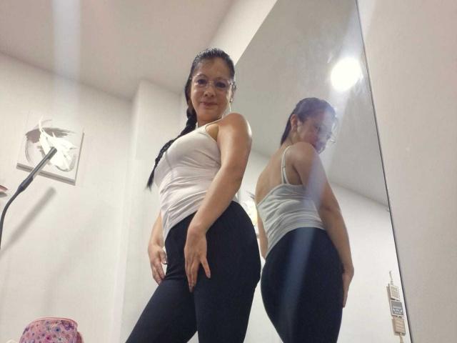 Low-angle photograph of MelissaBaud with a reflection. She's wearing a white tank top, black pants, and glasses. Her reflection shows her from the side. Bright light in the background.