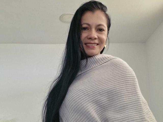 Melissa Baud, avec de longs cheveux noirs, souriant à la caméra, portant un pull blanc en tricot. Elle s'est légèrement tournée sur le côté, mettant en valeur sa silhouette élancée et sa petite poitrine. Fond clair et minimaliste.