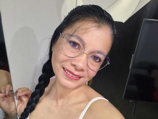 Melissa Baud, con i capelli neri intrecciati, occhiali chiari e canotta bianca, sorride con il rossetto rosso, tenendo in mano qualcosa di trasparente. con una cinghia al polso della mano destra. Illuminazione intensa, selfie informale.