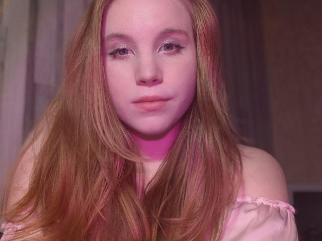 Close-up photo of SylvieOleksiak, a fair-skinned woman with long, reddish-brown hair, wearing a pink off-shoulder top. She has pink-tinted makeup, gazing directly at the camera with a slightly seductive expression.