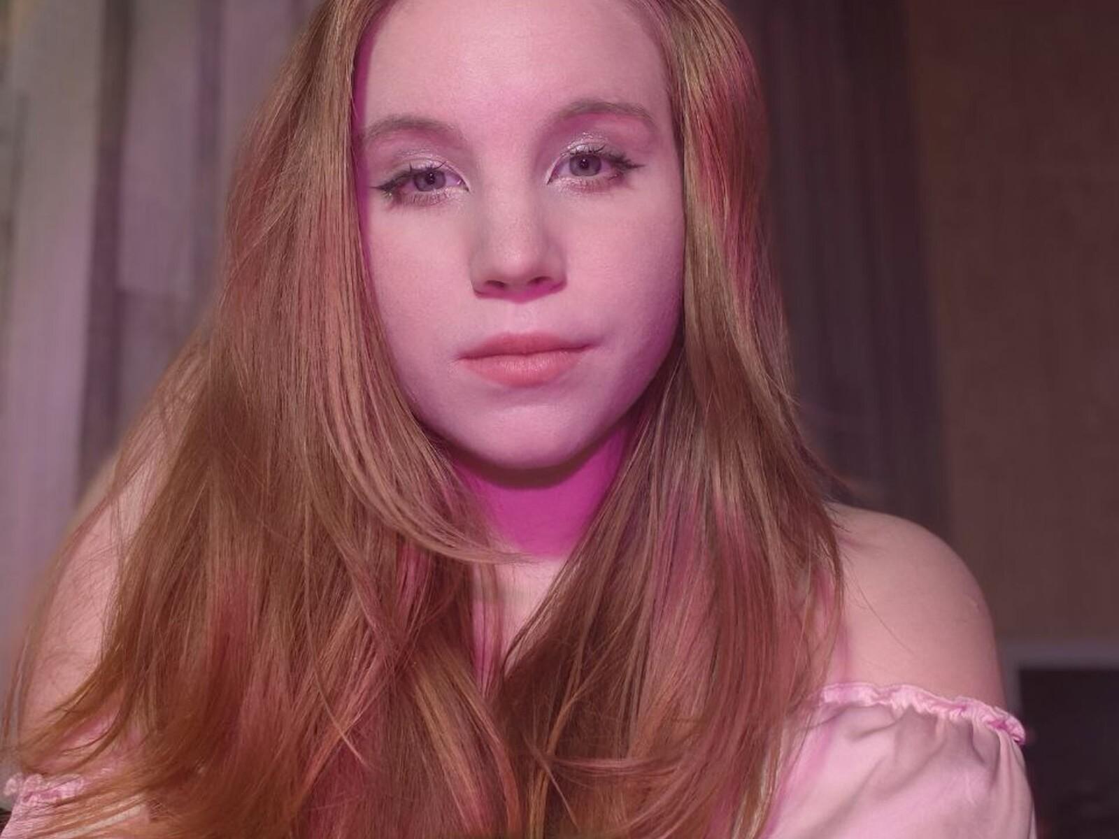Close-up photo of SylvieOleksiak, a fair-skinned woman with long, reddish-brown hair, wearing a pink off-shoulder top. She has pink-tinted makeup, gazing directly at the camera with a slightly seductive expression.