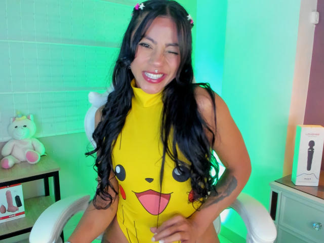 Amberv, una mujer de curvas con cabello largo y negro, guiña un ojo mientras está sentada en una silla blanca, vestida con un mono amarillo de Pikachu. Iluminación verde y blanca, unicornio de peluche a la izquierda, juguete sexual a la derecha..