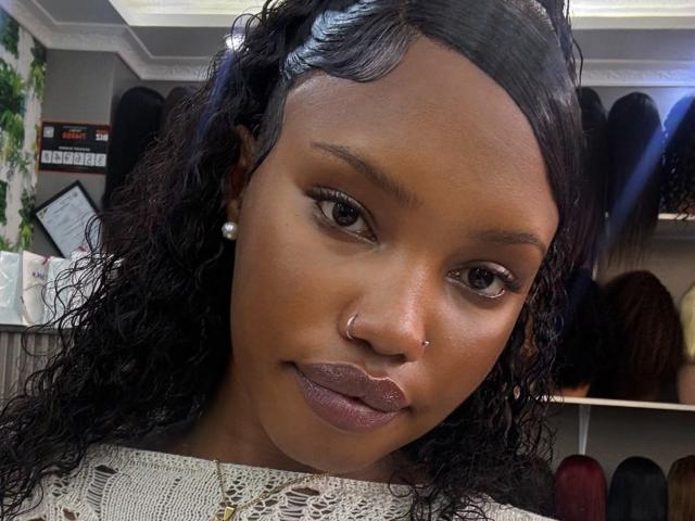 Fotografía de primer plano de LipaSianax, una atractiva mujer negra con cabello rizado, que lleva una blusa blanca de encaje, aretes de perlas y anillo para la nariz. Ella tiene labios carnosos y mira directamente a la cámara con una sonrisa sutil. El fondo muestra un entorno de oficina desenfocado..