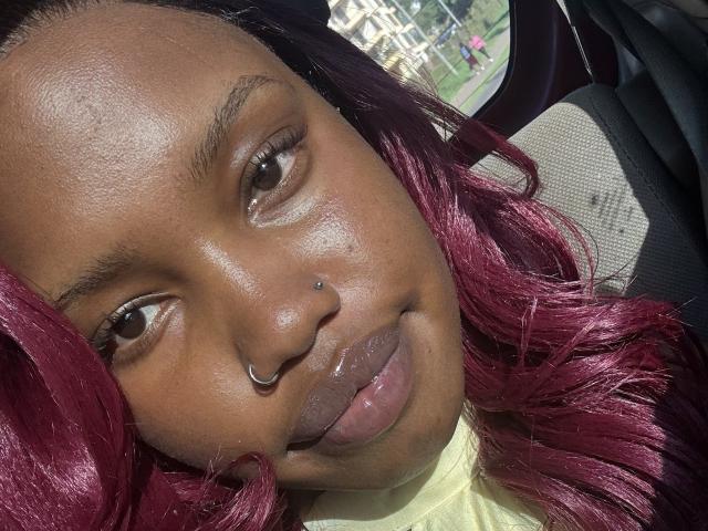 Close-up photo of LipaSianax with dark brown skin, glossy lips, and a silver nose ring. She has vibrant red hair, staring directly at the camera with a slight smile. Bright sunlight highlights her face.