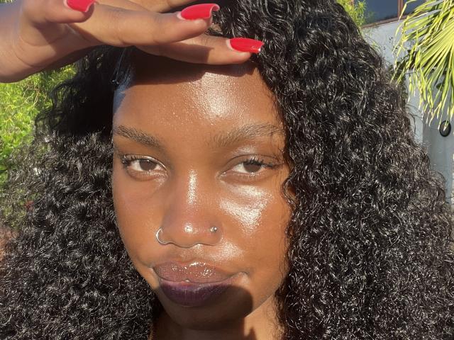 Primer plano de una LipaSianax con piel marrón oscuro brillante y cabello negro, grueso y rizado. Uñas rojas que tocan la cabeza. Rostro iluminado por el sol, brillante, con lápiz labial morado oscuro. La luz natural y vibrante resalta la textura..