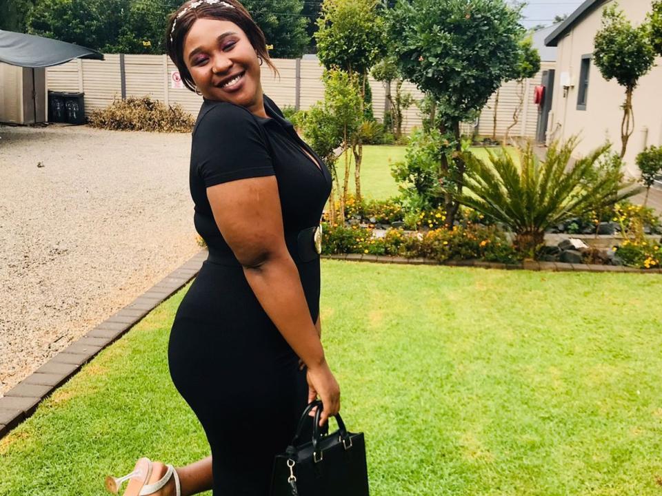 Fotografia de BadFatPussy69 ajoelhada em um gramado, vestindo um vestido preto justo, segurando uma bolsa preta, sorrindo. pele escura e corpo curvilíneo, ambiente de jardim ao ar livre..