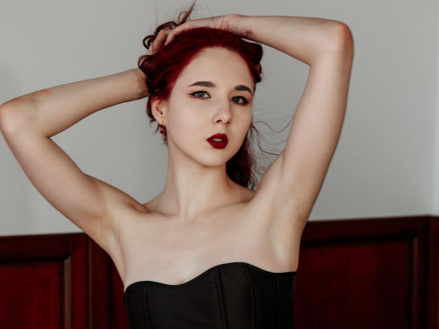 Photograph of MarieLoving with pale skin, dark red hair, and dark red lipstick. She's topless, wearing a black corset, with arms raised, hands in hair, exposing armpits, and a seductive expression.