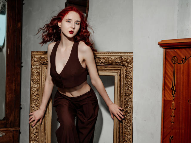 Fotografie von MarieLoving, einer blassen Frau mit roten Locken, die ein schwarzes Top mit tiefem V-Ausschnitt und eine lockere schwarze hosen, an einem aufwendig verzierten, goldenen Rahmen angelehnt, wirken verführerisch..