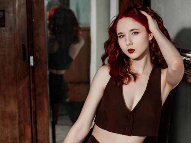 Fotografia di MarieLoving, una donna dalla pelle chiara e con i capelli rossi, che indossa una crop top nera con bottoni, con una mano tra i capelli, sguardo intenso. sguardo, rossetto rosso.
riflesso in uno specchio, sfondo con una porta di legno. Tensione sessuale..