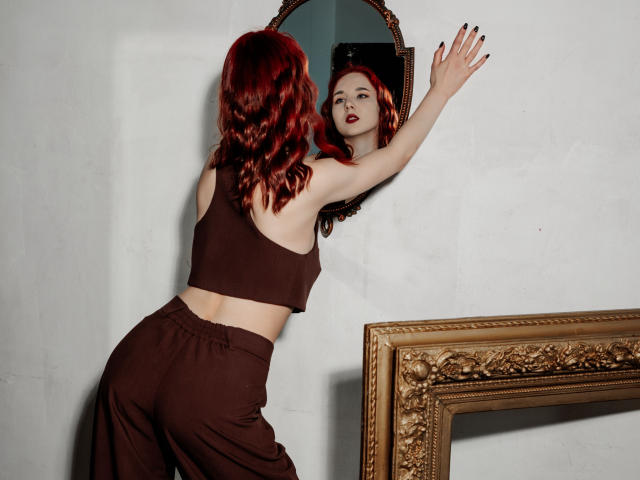 Marie, mit roten Haaren, einem schwarzen Crop-Top und hoch taillierten Hosen, lehnt sich an einen Spiegel, die Hand an der Wand, und blickt hinein. Reflexion, schattig, aufwendiger goldener Rahmen unten rechts, sexuelle Spannung..