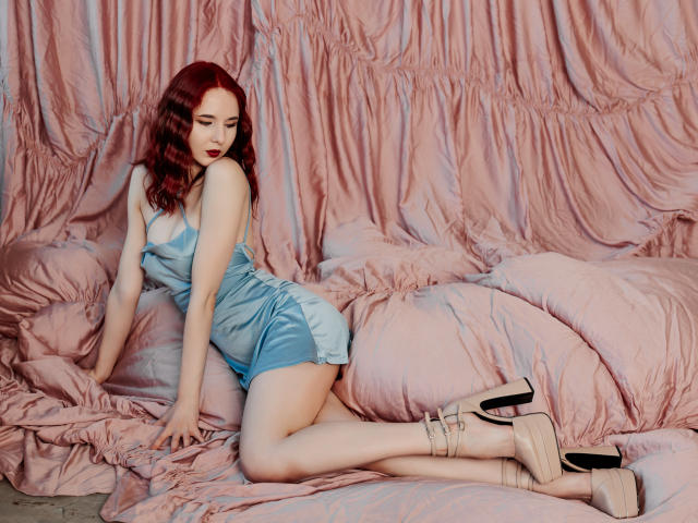 MarieLoving, pale-skinned, red-haired woman in tight blue satin dress, kneeling on pink ruffled bed, wearing beige wedge heels, eyes closed, seductive pose, soft pink background, sexy vibe.