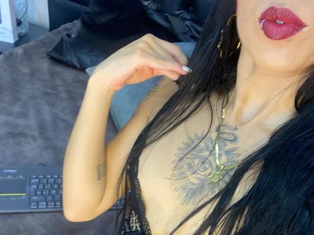 Close-up photo of MiaDeLune with black hair, full red lips, and a tattoo on her chest. She's wearing a beige top, touching her collarbone, next to a laptop. Fucking sexy.