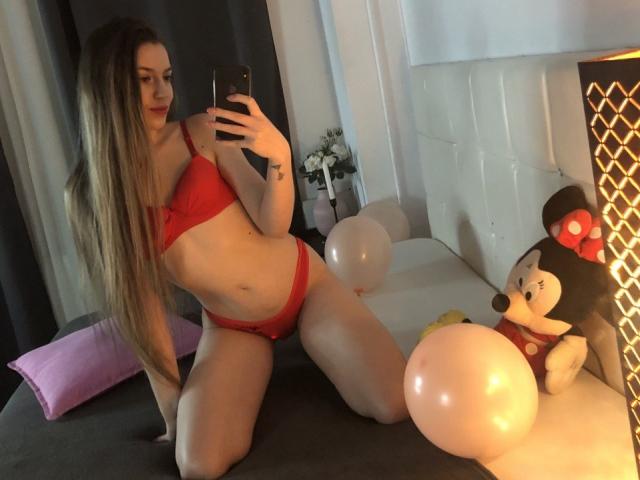 VelvetSpell knielt op een bed, gekleed in rode lingerie, en houdt een telefoon vast. Lang bruin haar, slank postuur, kleine borsten. Twee opgevulde poppen en ballonnen die oplichten in de buurt. Verleidelijke, uitdagende pose.