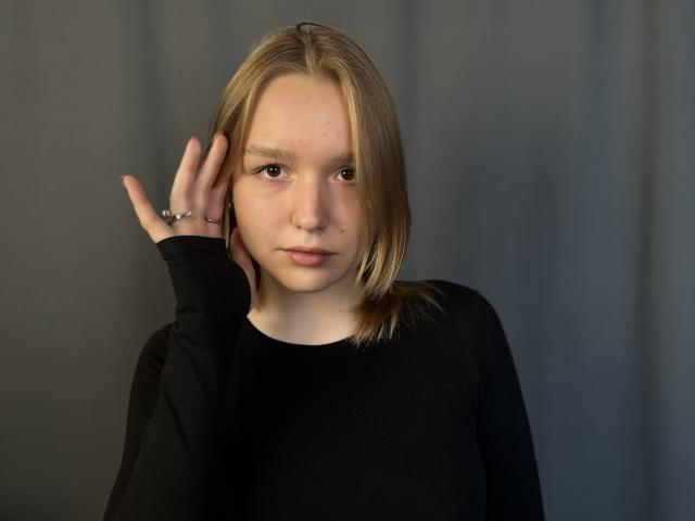 Photographie de Rima Pecinovsky, avec des cheveux brun clair, portant une chemise noire à manches longues, sa main touchant sa tête, prise en plongée. regard, expression neutre, lumière douce, légère ombre, fond gris, tension sexuelle subtile.