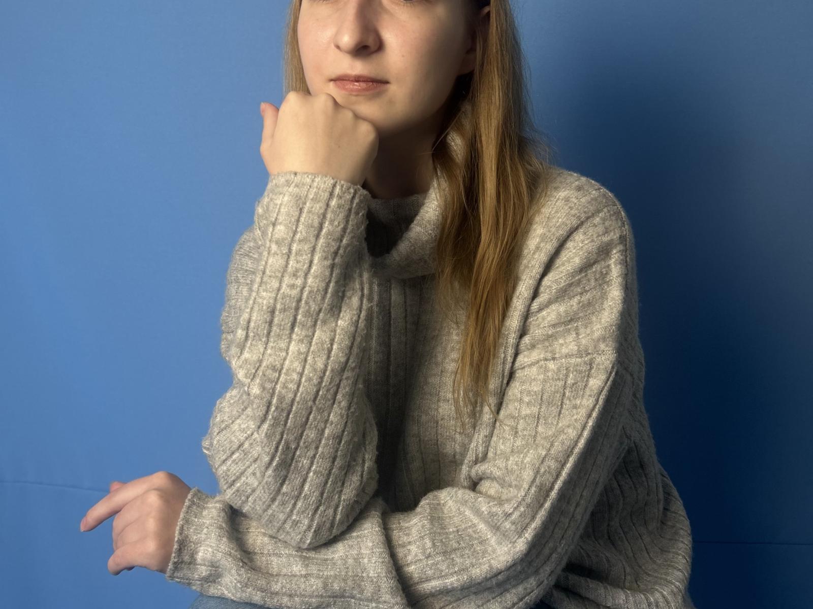 ShaniquaKrivanec, avec ses longs cheveux bruns, portant un pull gris côtelé, repose son menton sur sa main, regardant légèrement. vers le bas, sur un fond bleu vif. Les ombres douces rehaussent son expression contemplative..