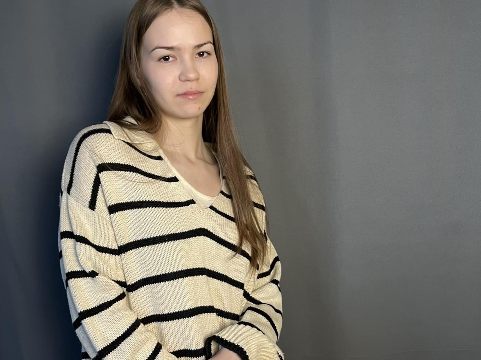 Fotografía de MarieBellas con cabello largo y castaño, vistiendo un suéter blanco con rayas negras, de corte holgado, de pie frente a un fondo oscuro. Fondo gris.
su expresión es neutra, con un ligero indicio de tensión sexual..