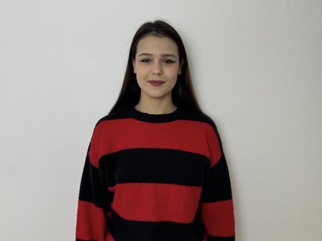 MandaSecora steht vor einer weißen Wand, trägt einen rot-schwarz gestreiften Pullover und hat langes, schwarzes Haar. Ihr Gesichtsausdruck ist neutral. Ausdruck, leichtes Lächeln, weiches Licht, lockere Pose, keine sichtbaren Details im Hintergrund.