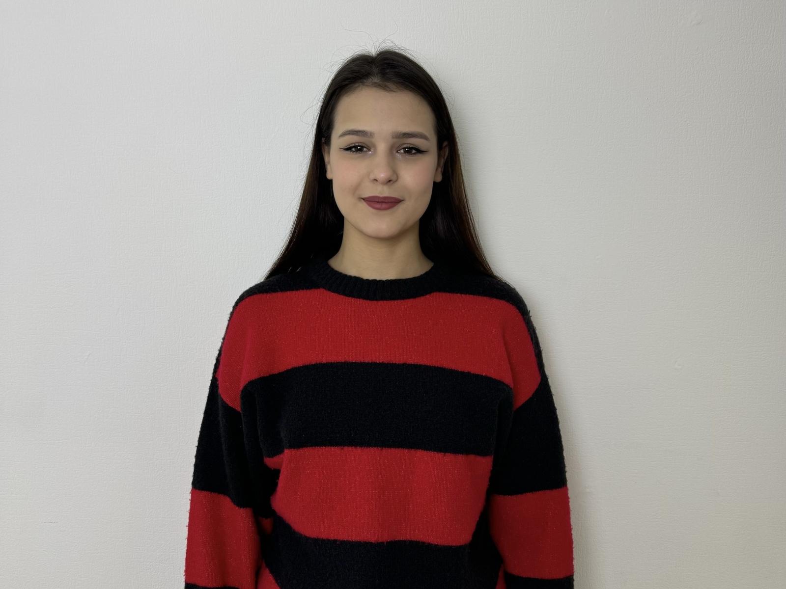 MandaSecora, de pie frente a una pared blanca, vestida con un suéter a rayas rojas y negras, con cabello largo y negro, expresión neutra. expresión, ligera sonrisa, iluminación suave, pose informal, sin detalles visibles del fondo.