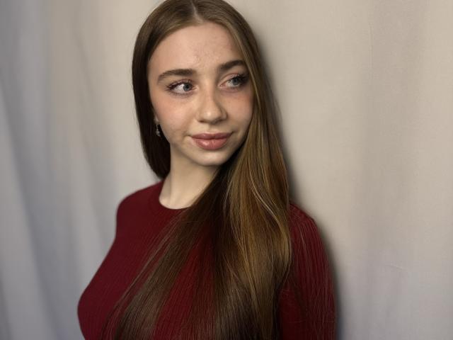FelicitasDeroy with long brown hair, wearing a red sweater, has a cute smile, small tits, and light skin, looking to the side with a subtle, confident expression.