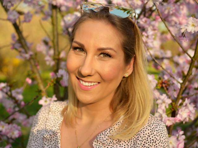 Photo en gros plan de Simone, souriante, avec des yeux brillants, des cheveux blonds et un haut blanc à pois. Des lunettes de soleil reposent sur sa tête. Arrière-plan : des cerisiers en fleurs roses, flous. Éclairage vif et ensoleillé.