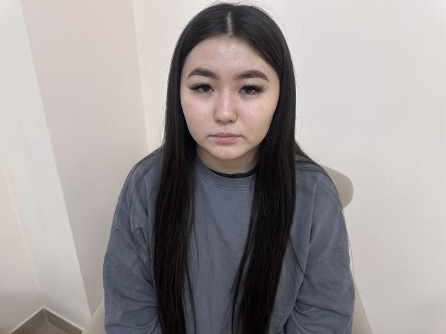 Foto van Emily Johnsone met lang zwart haar, die een grijze trui draagt en tegen een witte muur zit. Ze heeft een neutrale uitdrukking, een lichte blos en maakt direct oogcontact. Een serieus, intens blik..