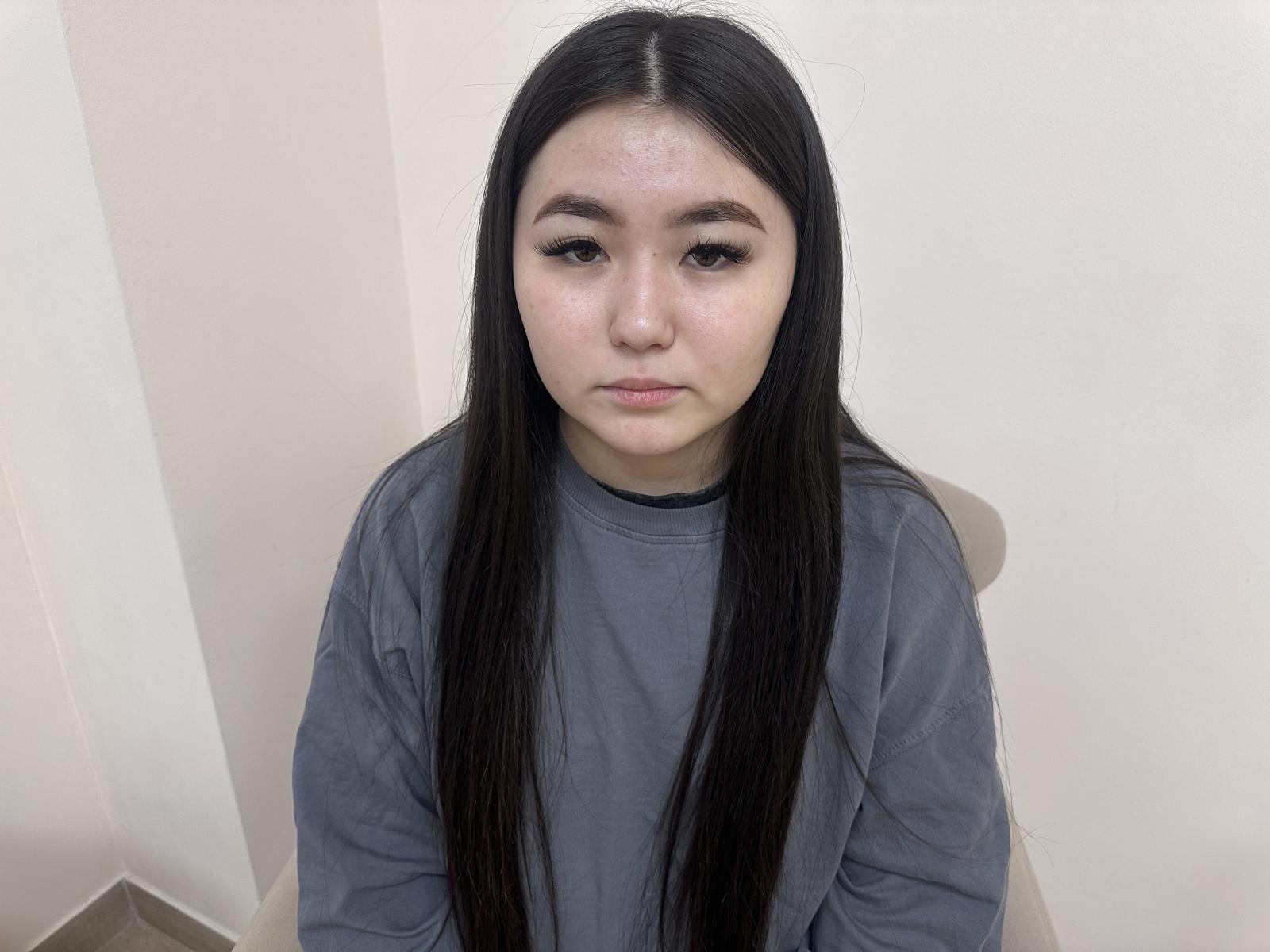 Foto van Emily Johnsone met lang zwart haar, die een grijze trui draagt en tegen een witte muur zit. Ze heeft een neutrale uitdrukking, een lichte blos en maakt direct oogcontact. Een serieus, intens blik..