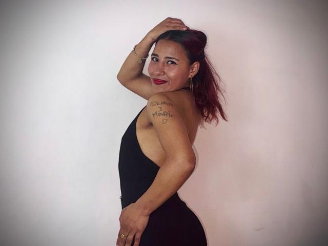 JenSthifler with medium skin tone, red lipstick, and black dress, standing sideways, hand in hair, tattoo on left shoulder, sultry smile, raised arm, white background, sexy pose, confident.