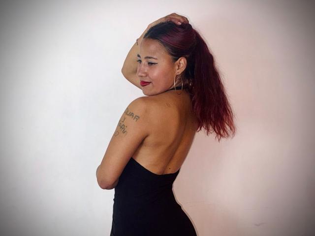 Fotografía de JenSthifler con cabello castaño oscuro en una coleta, vestido negro sin mangas, tatuaje en el hombro izquierdo, mirando mirando por encima del hombro, con lápiz labial rojo. Fondo blanco, tensión sexual, pose segura..