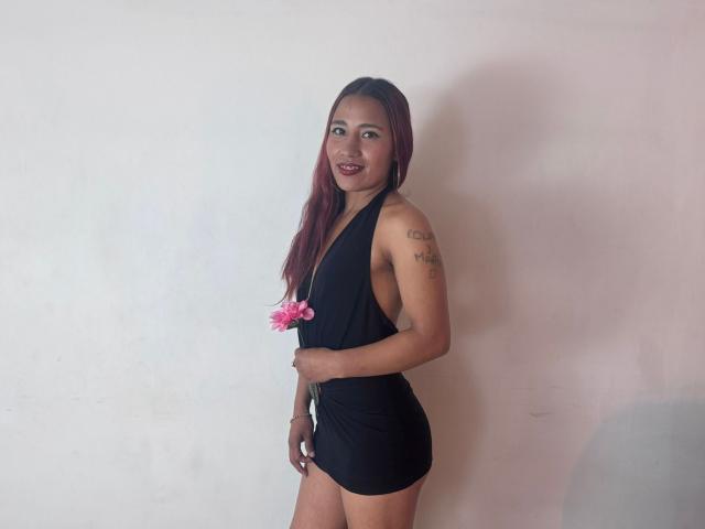 Photograph of JenSthifler standing against a plain white wall, wearing a black halter top and shorts, holding pink flowers, smiling, with a tattoo on her upper arm, long brown hair.