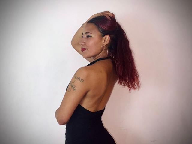 JenSthifler è in piedi rivolta a sinistra, con la schiena arcuata, un abito nero che rivela un tatuaggio sulla spalla, e una mano tra i capelli castani. Sfondo bianco. Labbra rosse, espressione sicura, leggero sorriso, posa sensuale, visibile una parte del seno..