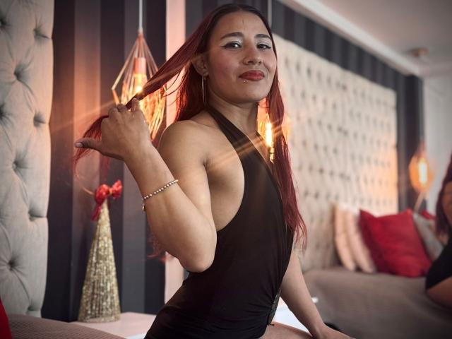 JenSthifler, à la peau brune, avec de longs cheveux roux, portant un haut noir à encolure dégagée, tenant ses cheveux dans sa main droite, avec un sourire séduisant, regardant la caméra, pièce faiblement éclairée, décoration moderne, éclairage doux.