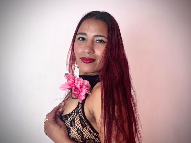 Fotografia di JenSthifler con lunghi capelli lisci e rosso scuro, che indossa una maglia nera a rete e una collana di fiori rosa. Sorridendo, con rossetto rosso, mentre si tiene il petto, sfondo bianco. Molto attraente..