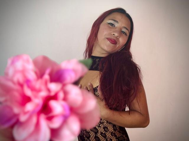 JenSthifler with long red hair, wearing a black lace dress, holding a blurred pink flower in the foreground. She gazes at the camera with a slight, seductive smile.