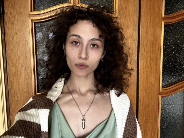 AlyceErway with curly brown hair, light skin, wearing a green top and brown-striped cardigan, necklace with a teardrop pendant, standing in front of wooden doors.